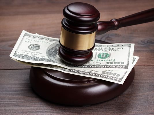 A wooden judge's gavel resting on a stack of $100 bills on a dark wood surface