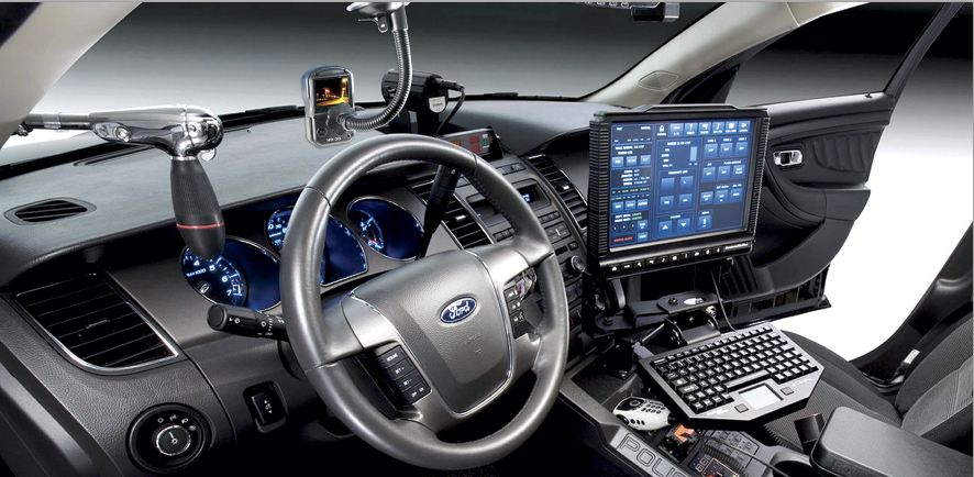 The front cabin of a Ford patrol car featuring a specialized tech setup: a large digital display with various control panels, a compact keyboard, and secondary monitors mounted to the windshield and dashboard