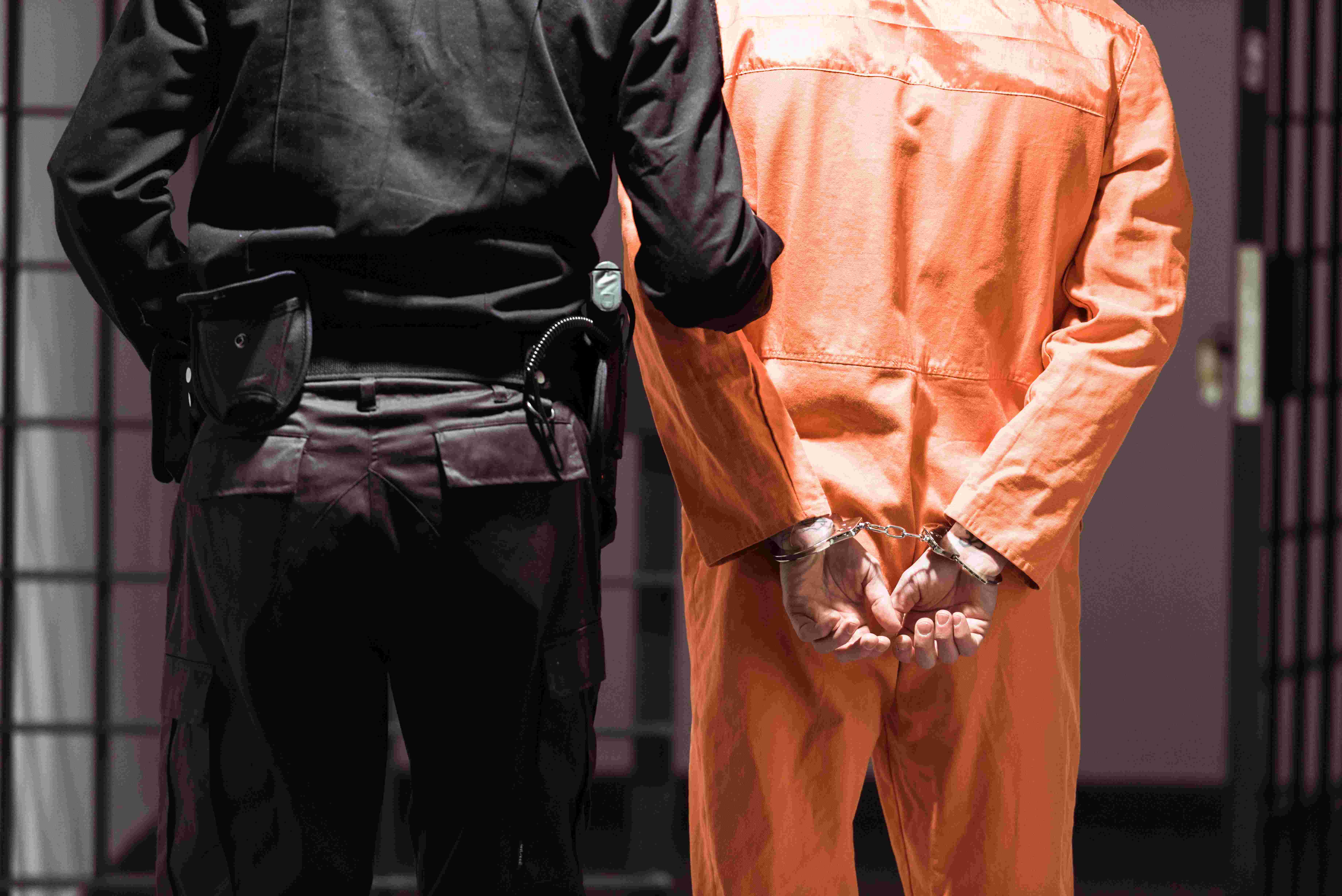 Back view of a law enforcement officer in a black uniform escorting a person in an orange jumpsuit and handcuffs