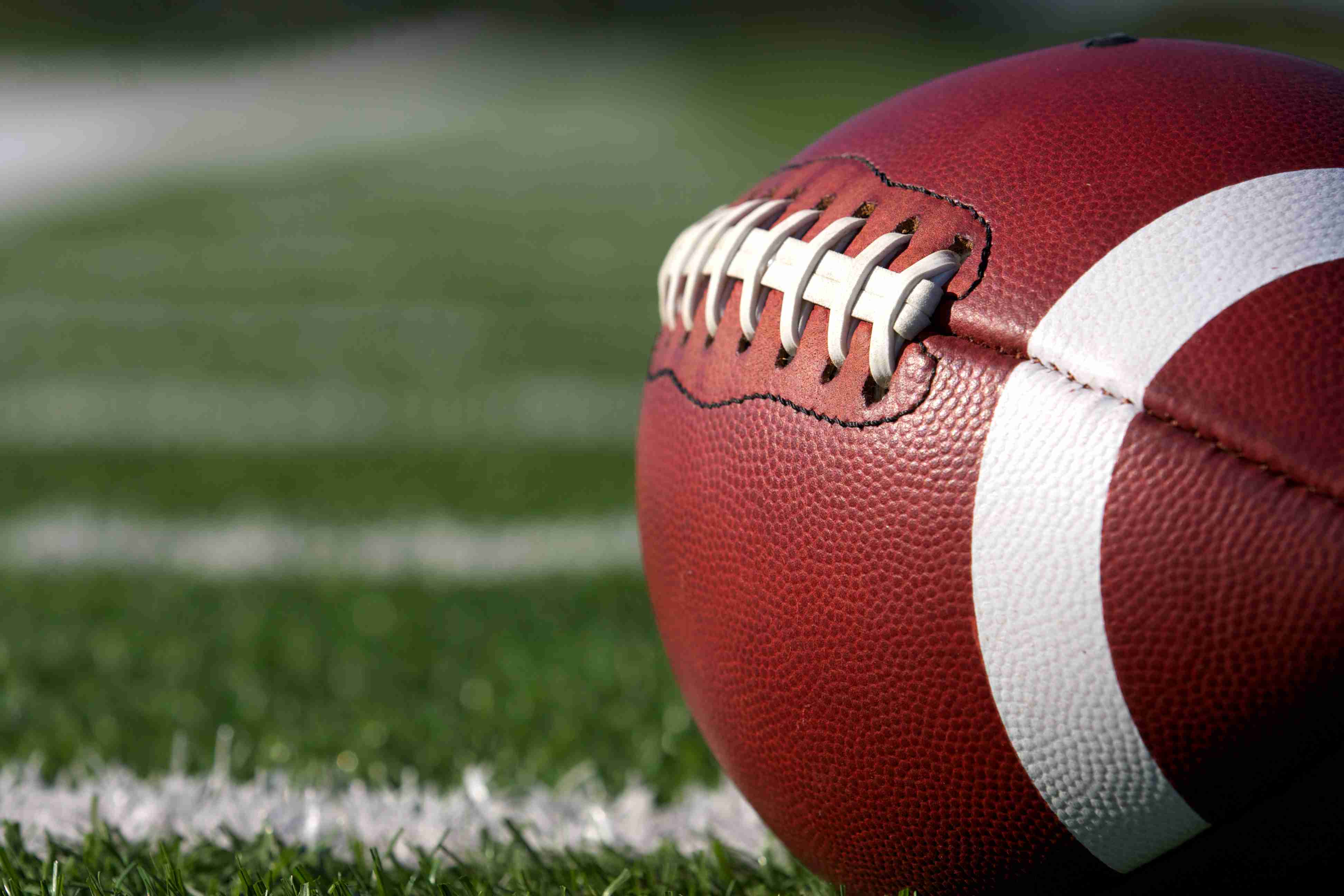 A close-up of a brown football with white laces and stripes resting on a green turf field with white yard lines