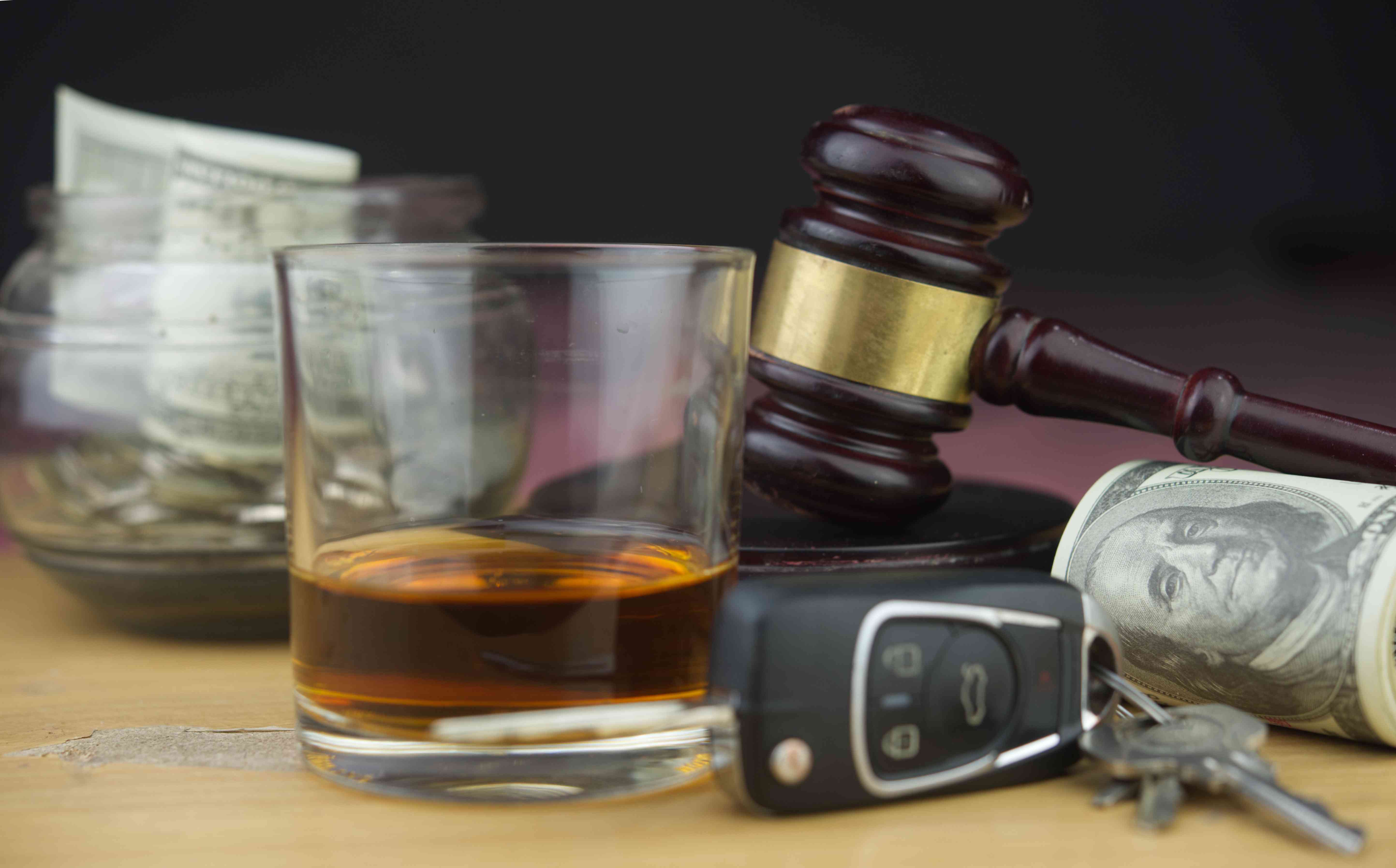 A close-up of a glass of whiskey, car keys, a wooden judge's gavel, and rolls of hundred-dollar bills on a wooden surface