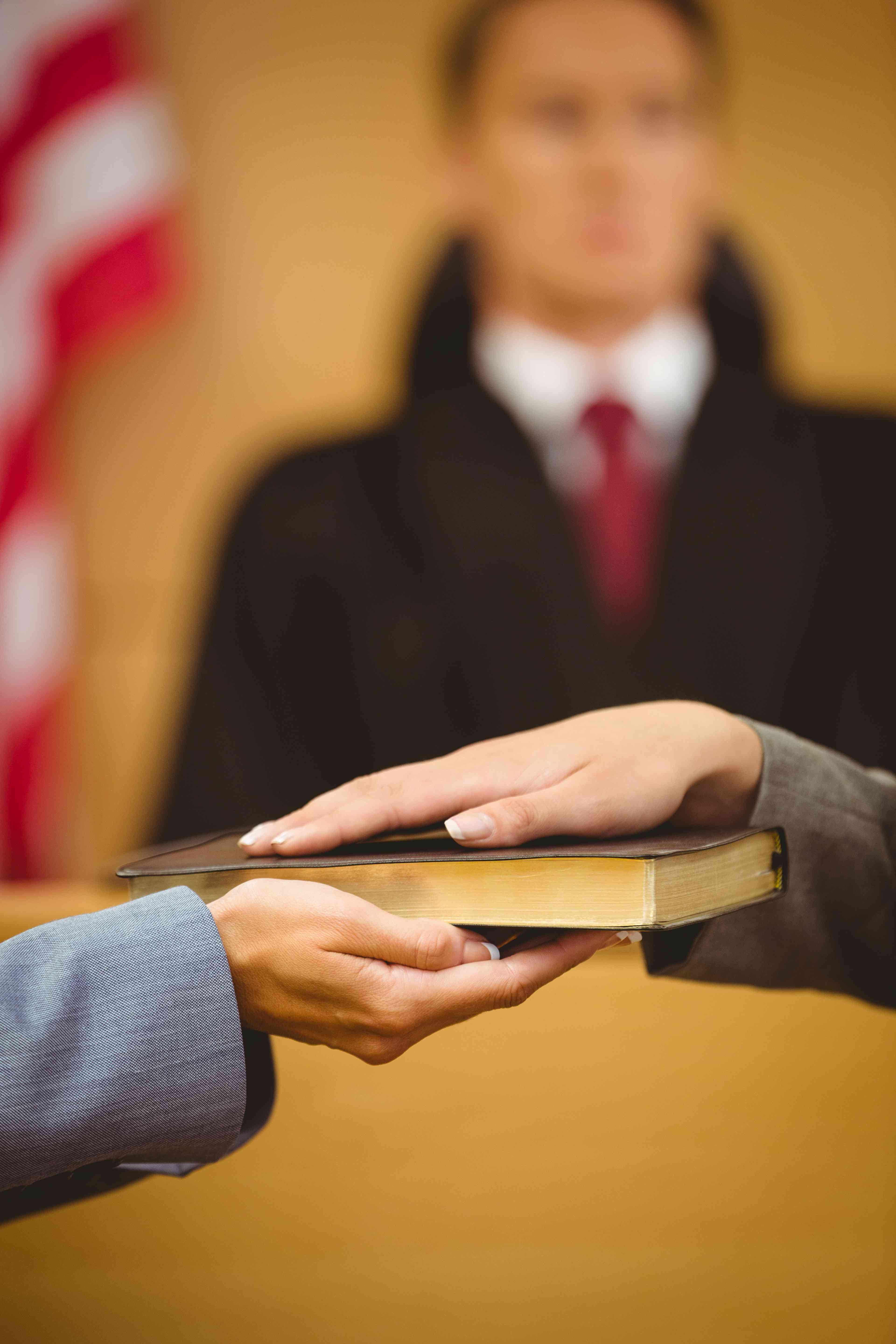 Close-up of a person placing their hand on a book to swear an oath in a courtroom, with a judge blurred in the background